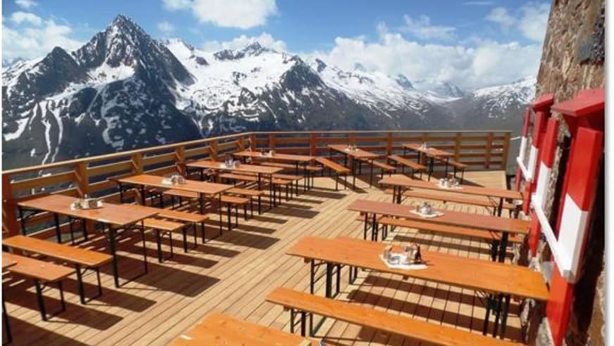 Sonnenterrasse der Breslauer Hütte mit Holzdielen und Biertischgarnituren vor schneebedecktem Bergpanorama der Ötztaler 
