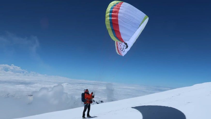 Bergführerin Elisabeth Seibt hält einen bunten Gleitschirm über sich auf verschneitem Gipfelplateau über einer weißen Wolkendecke.
