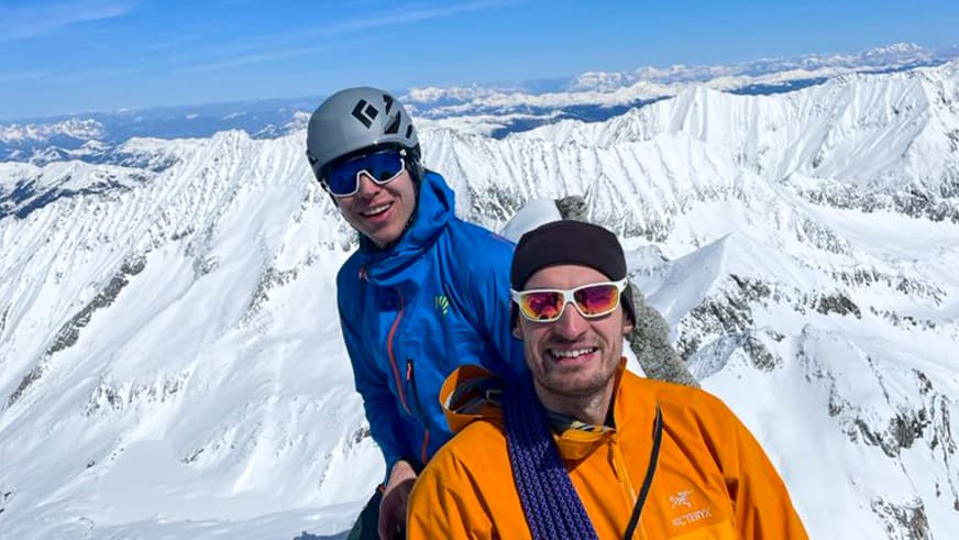 Zwei Personen posieren für Gipfelfoto, darunter Bergführer Alexander Reisenhofer vor schneebedeckten Bergen.