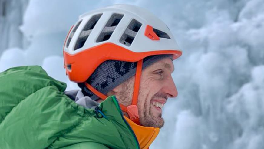 Bergführer Alexander Reisenhofer zeigt sich im Profil mit weißem Kletterhelm und grüner Softshell-Jacke vor schneebedeckten Bergen.