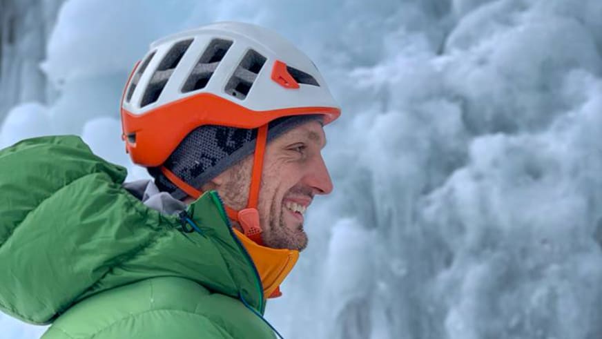 Bergführer Alexander Reisenhofer lächelt mit weißem Kletterhelm vor schneebedeckter Berglandschaft.