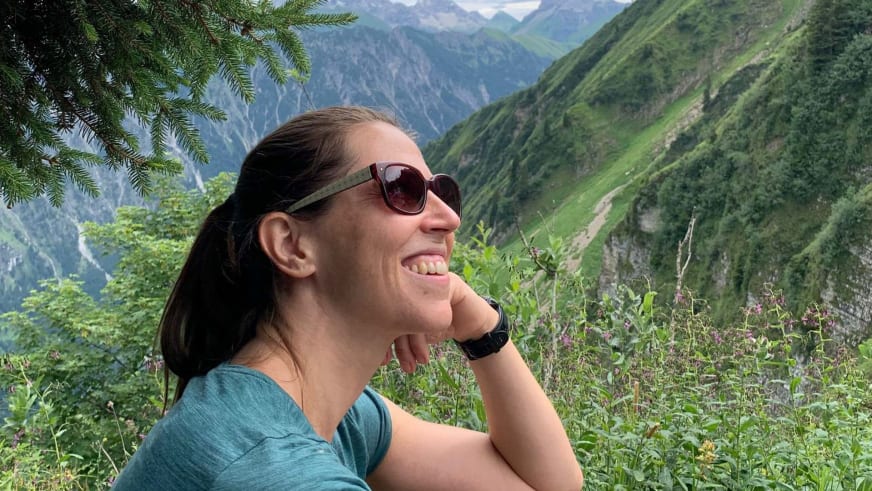 Bergwanderführerin Charlotte Malycha mit Sonnenbrille und türkisblauem T-Shirt vor einem tiefen Tal mit steilen grünen Berghängen.