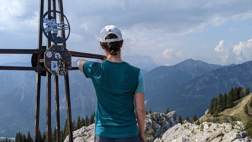 Am metallenen Gipfelkreuz zeigt Bergwanderführerin Charlotte Malycha mit ausgestrecktem Arm in die alpine Bergkette.