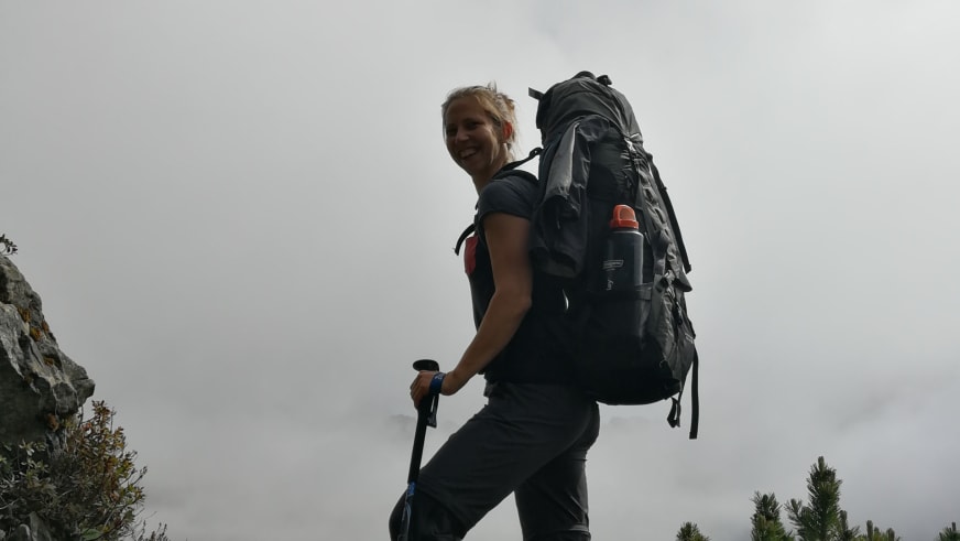 Bergwanderführerin Charlotte Malycha steht lächelnd mit schwarzem Trekkingrucksack und Wanderstock vor bewölktem Himmel.