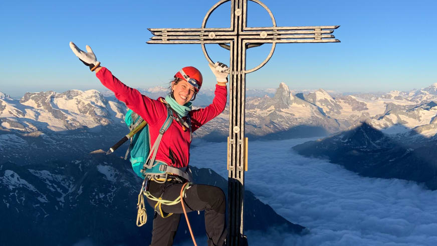 Bergwanderführerin Charlotte Malycha streckt beide Arme jubelnd am hölzernen Gipfelkreuz hoch, trägt rote Jacke und Eispickel.