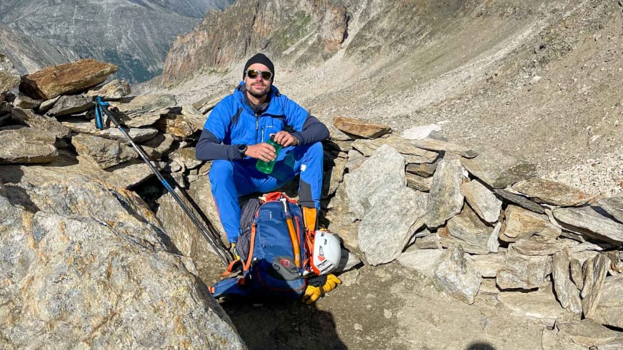 Bergführer Tim Lenz pausiert in blauer Daunenjacke auf Felsblock mit grüner Trinkflasche und Rucksack.