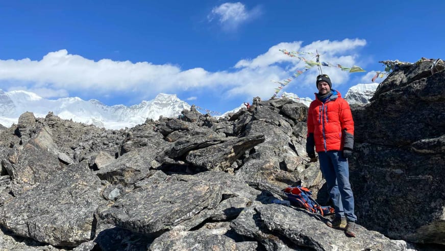 Bergführer Tim Lenz steht in roter Daunenjacke auf einem Gipfel zwischen Felsblöcken mit Gebetsfahnen.