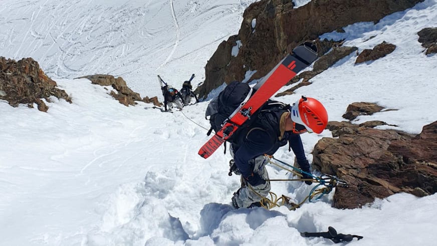 Bergführer Tim Lenz steigt mit Eisgeräten durch steilen, verschneiten Felshang, Skier am Rucksack befestigt.