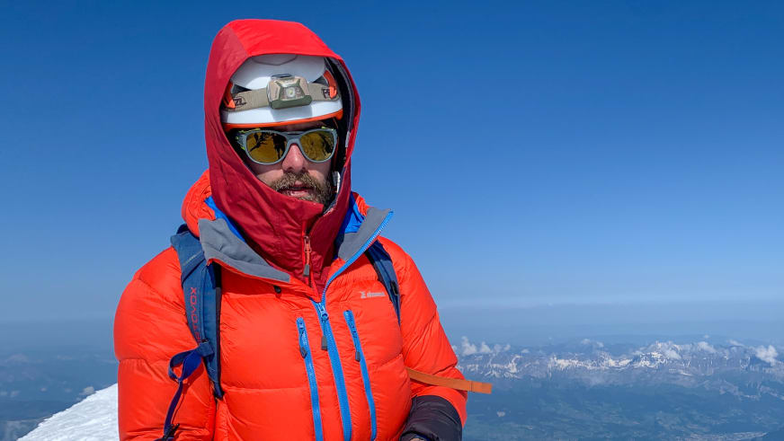 Portrait von Bergführer Tim Lenz in oranger Daunenjacke mit Helm und Stirnlampe vor Bergkette.