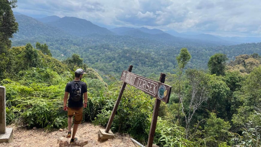 Bergführer Tim Lenz steht vor hölzernem Wegweiser 'BUKIT TERISEK' mit Blick über Dschungellandschaft.