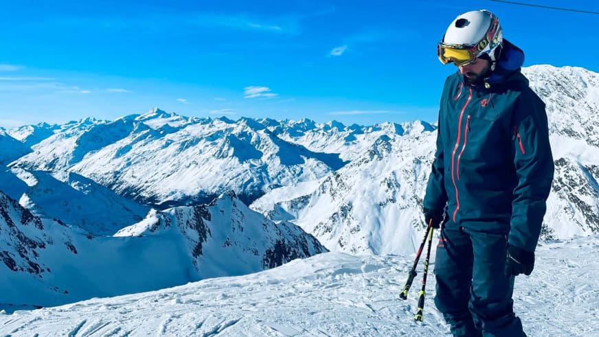 Bergführer Tim Lenz steht mit weißem Skihelm und Skistöcken auf verschneitem Gipfel vor Bergpanorama.