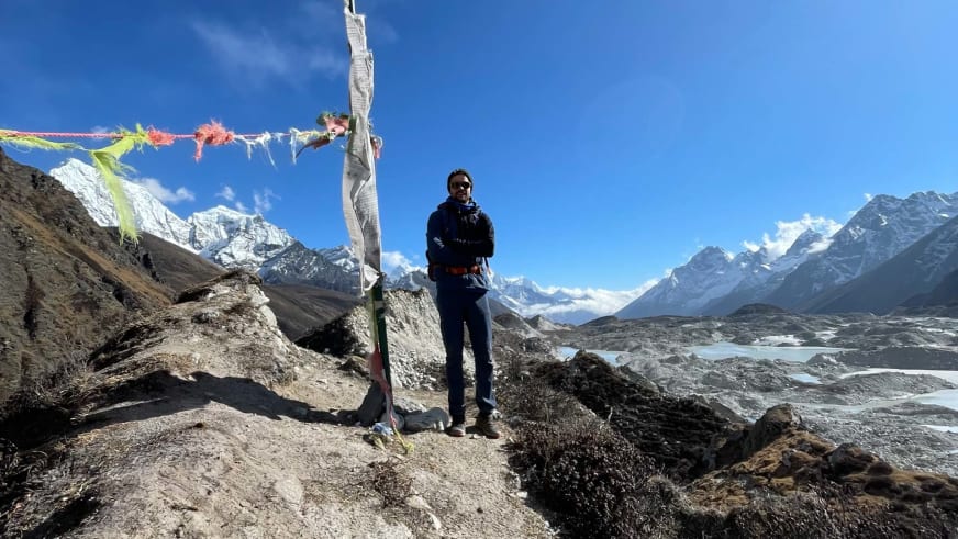 Bergführer Tim Lenz steht neben bunten Gebetsfahnen mit Blick auf Gletschersee und schneebedeckte Gipfel.