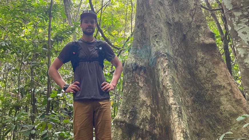 Bergführer Tim Lenz steht mit Händen in Hüften vor großem Baumstamm im tropischen Wald.