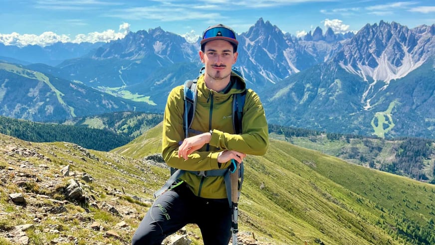 Bergwanderführer Tobias Storz sitzt mit gelbem Cap und Softshelljacke auf steinigem Berghang vor Berglandschaft.