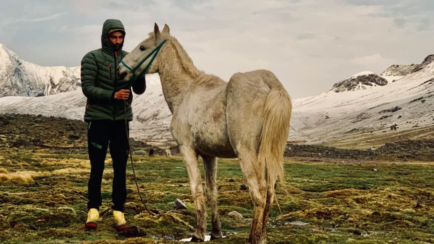 In grüner Winterjacke steht Bergwanderführer Tobias Storz neben hellem Pferd in verschneiter Hochgebirgslandschaft.