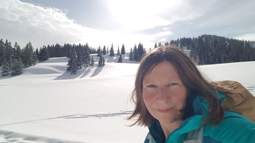 Bergwanderführerin Anna Schärtel lächelt in türkiser Winterjacke vor verschneiter Landschaft mit bewaldeten Hügeln.
