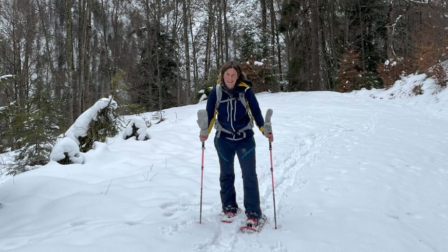 Auf verschneitem Waldweg steht Bergwanderführerin Anna Schärtel lächelnd mit roten Schneeschuhen und Stöcken in den Händen.