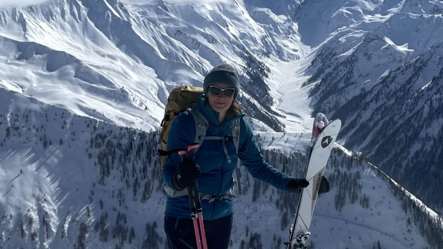 Anna Schärtel trägt Skier über der Schulter und steht als Bergwanderführerin in verschneitem Hochtal mit Nadelwald.
