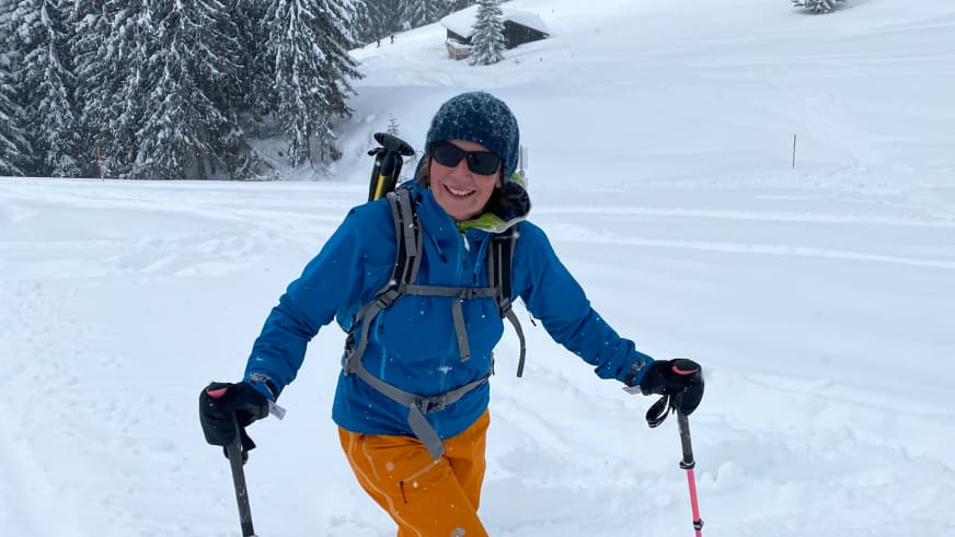 In verschneiter Winterlandschaft lächelt Bergwanderführerin Anna Schärtel in blauer Jacke mit Wanderstöcken und grauem Rucksack.