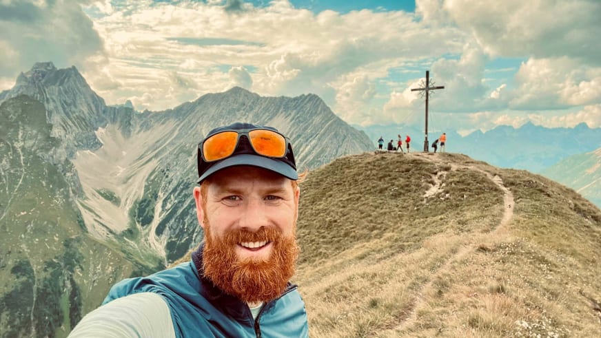 Selfie von Bergwanderführer Dennis Enders mit orangener Gletscherbrille vor alpiner Bergkulisse mit Gipfelkreuz.