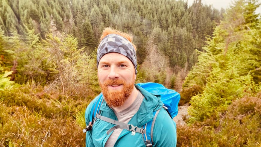 Portrait von Bergwanderführer Dennis Enders mit rotem Vollbart und türkiser Wanderjacke vor Nadelwald.