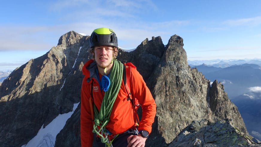 Bergführer Florian Storkenmaier trägt orange Softshelljacke und grünen Helm auf felsigem Gipfel.