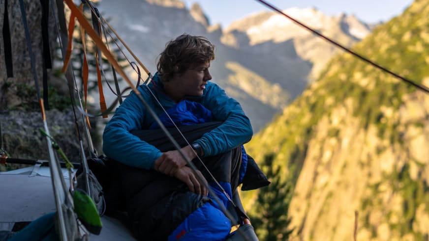 Bergführer Florian Storkenmaier sitzt in blauer Daunenjacke auf Zelunterlage am steilen Felshang.