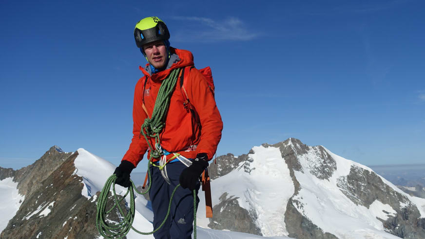 Am schneebedeckten Gipfel steht Bergführer Florian Storkenmaier mit gelbem Helm und grünem Kletterseil.