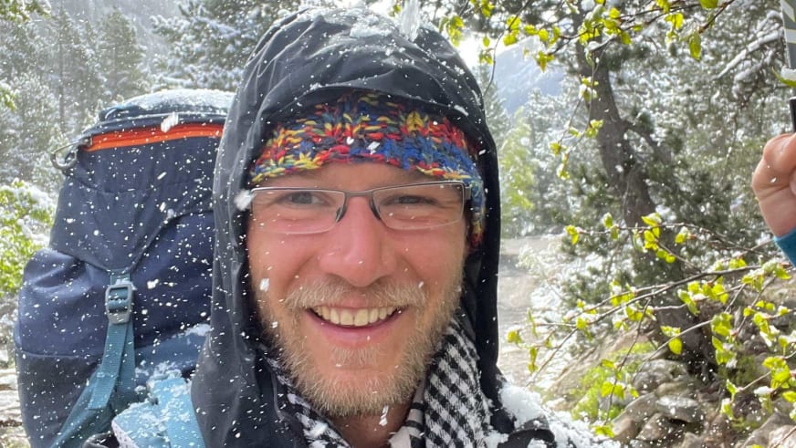 Portrait von Bergwanderführer Johannes Geipel mit Brille und bunter Strickmütze bei dichtem Schneefall in verschneiter Berglandschaft.