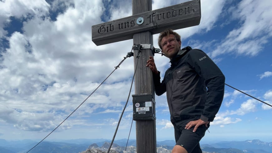 Bergwanderführer Johannes Geipel steht am Holzkreuz eines Berggipfels und berührt den Kreuzpfosten mit der rechten Hand.