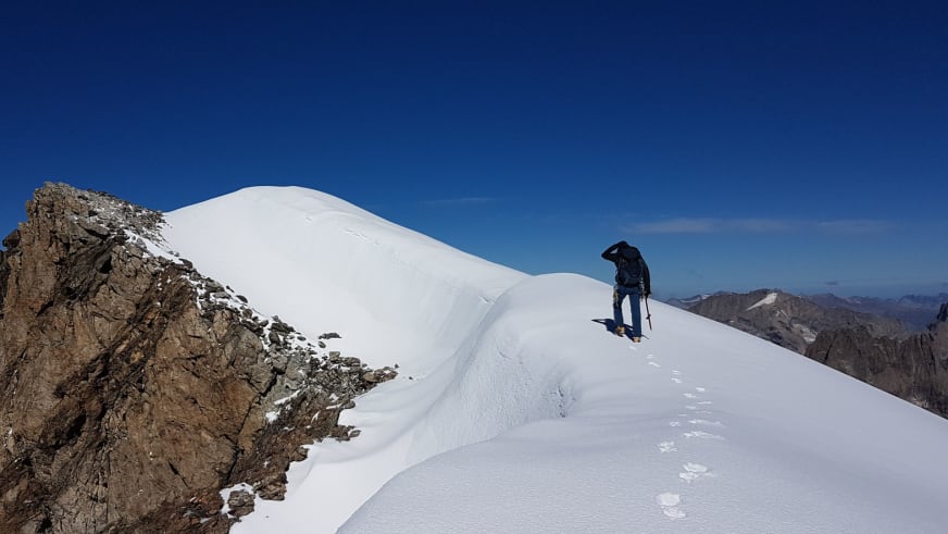 Bergwanderführer Johannes Geipel steht mit Steigeisen auf verschneitem Grat zwischen steiler Felswand und weißer Firnfläche bei klarem Himmel.