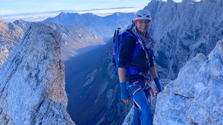 Bergwanderführer Johannes Geipel in weißem Kletterhelm und blauer Softshell-Jacke steht auf hellem Kalkfelsgrat mit Bergpanorama im Hintergrund.