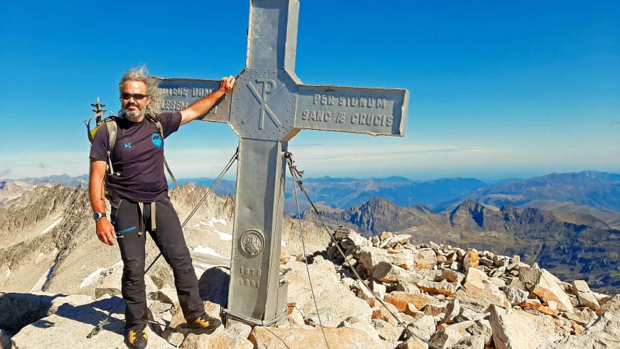 Bergwanderführer Nacho Carreras Codina legt die Hand auf das Gipfelkreuz mit weiter Fernsicht über die Berglandschaft.