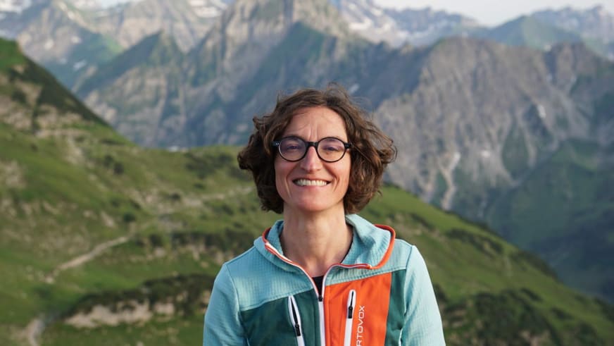 Bergwanderführerin Marie Nimsgern mit lockigen Haaren und runder Brille vor alpiner Berglandschaft mit schneebedeckten Gipfeln im Hintergrund.