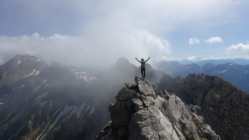 Mit erhobenen Armen jubelt Bergwanderführerin Marie Nimsgern auf einem hellen Kalksteingipfel vor weitem Alpenpanorama.
