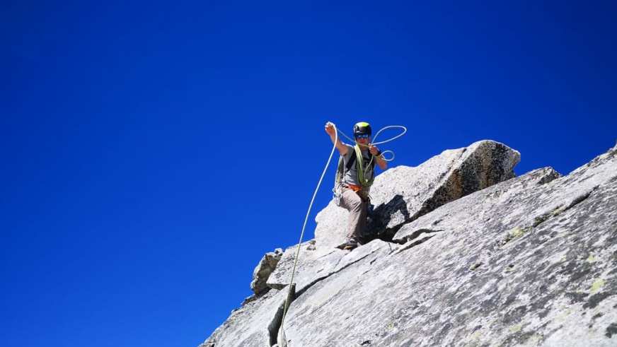 Bergführer Stefan Scheiblauer sitzt mit grünem Seil und gelbem Helm auf steilem Granithang vor blauem Himmel.