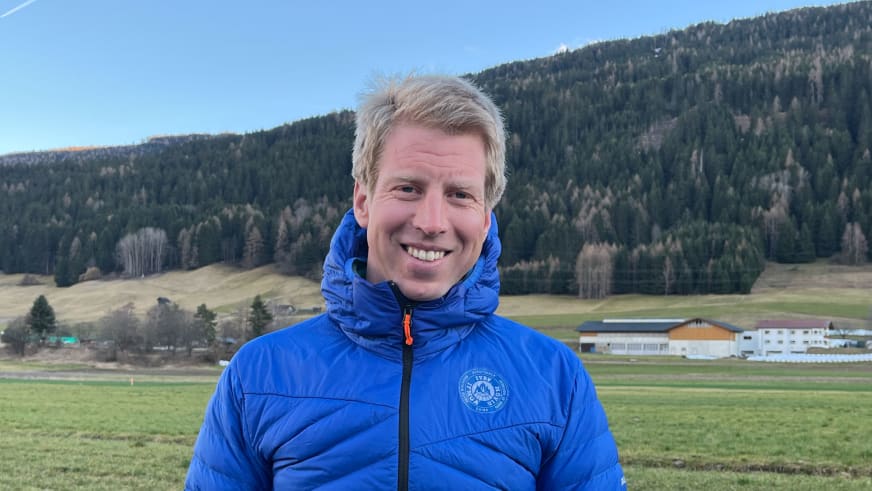 Bergführer Stefan Scheiblauer in blauer Daunenjacke lächelt vor alpiner Landschaft mit bewaldeten Hängen und grünen Wiesen.