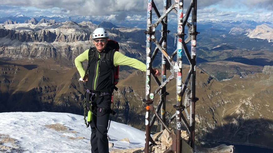 Bergwanderführer Gerald Römpp mit weißem Helm und Klettergurt steht am metallenen Gipfelkreuz vor weitem Bergpanorama.