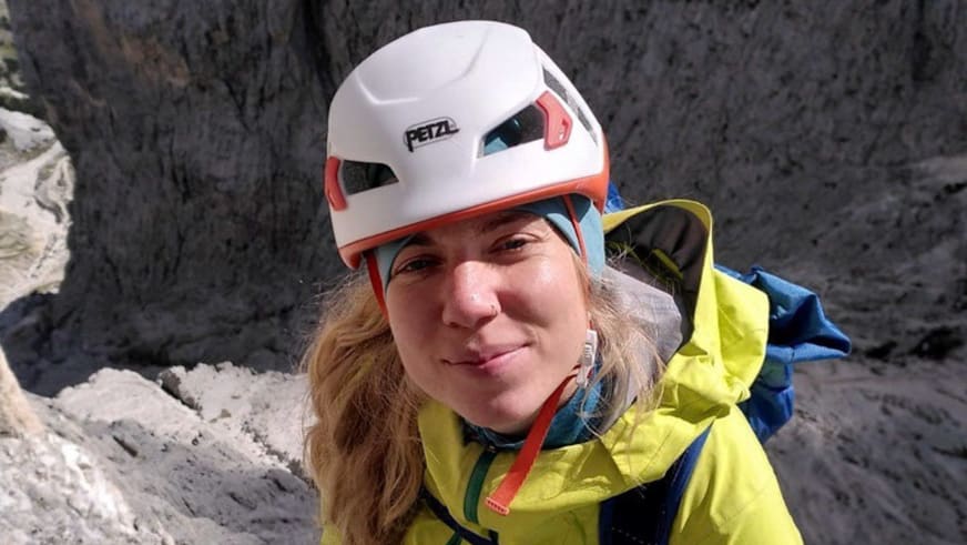 Bergwanderführerin Sophie Sasse trägt einen weißen Petzl-Kletterhelm und gelbe Outdoorjacke vor einer dunklen Felswand.