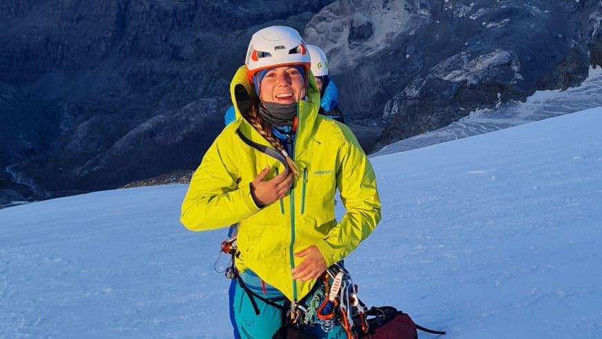 Bergwanderführerin Sophie Sasse trägt gelbe Softshell-Jacke und weißen Helm auf schneebedecktem Gelände mit Bergkulisse.