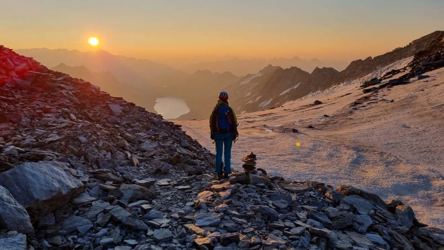 Bergwanderführerin Sophie Sasse steht in blauer Jacke auf steinigem Bergplateau und blickt bei Sonnenaufgang über mehrere Bergketten.