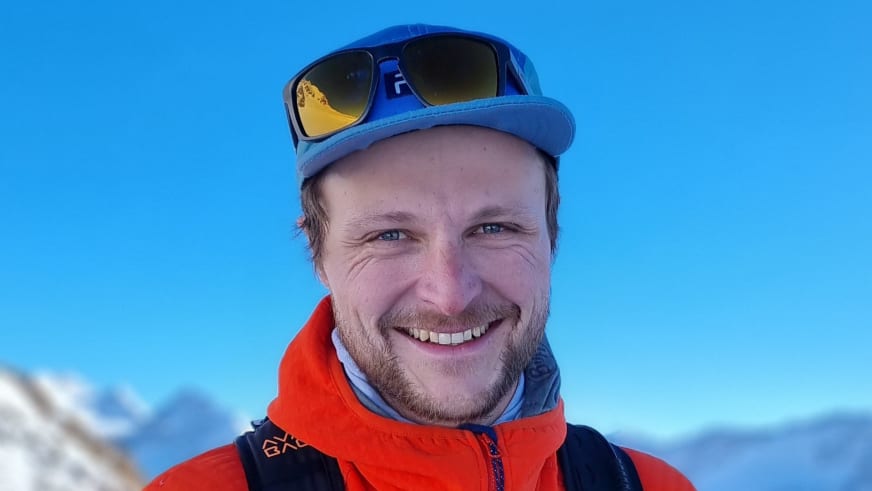 Bergführer Tobias Kostenbader lächelt vor verschneiter Bergkulisse, trägt orange-schwarze Winterjacke und blaue Mütze.