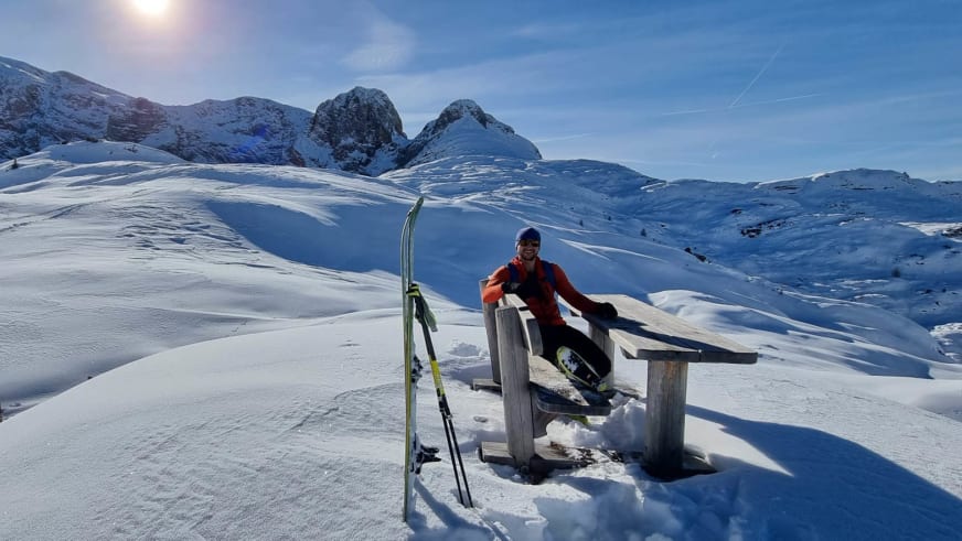 Bergführer Tobias Kostenbader sitzt in orangeroter Winterjacke auf einer Holzbank in verschneiter Berglandschaft.