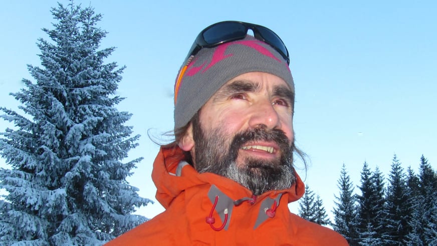 Portrait von Bergwanderführer Karsten Clauß mit grauer Strickmütze und orange-grauer Winterjacke vor verschneiten Nadelbäumen.