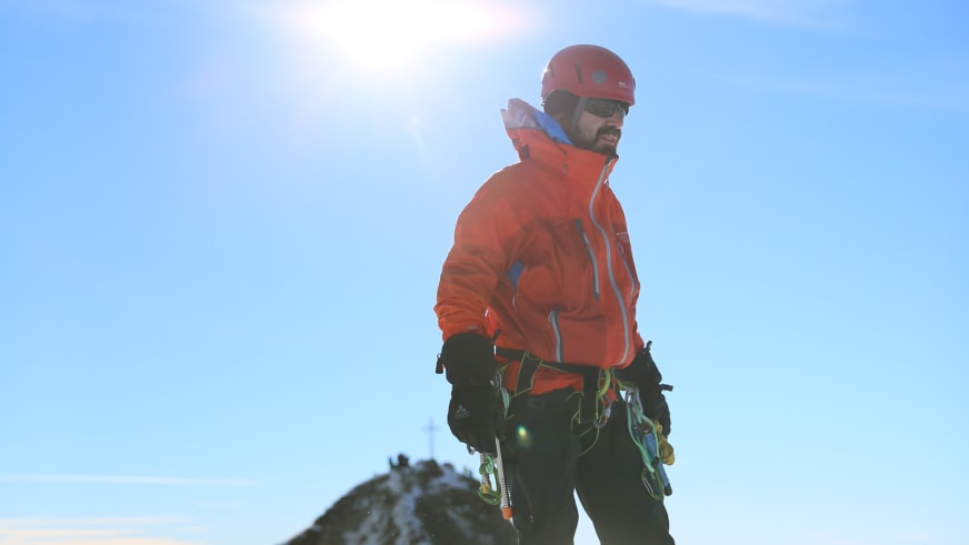 Mit rotem Kletterhelm und oranger Softshelljacke steht Bergwanderführer Karsten Clauß vor blauem Himmel und Felsgrat.