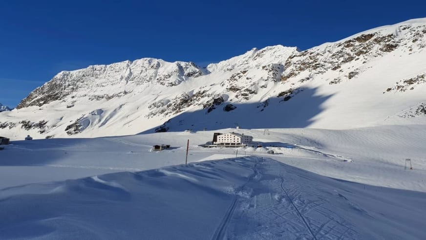 Das weiße Gebäude des Berggasthof Piz Buin steht auf einem schneebedeckten Plateau mit präparierten Skipisten rundherum.