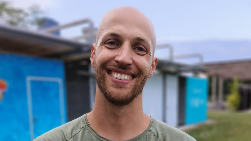 Bergführer Alexander Berger lächelt in die Kamera, trägt olivgrünes Langarmshirt vor unscharfem Gebäudehintergrund.