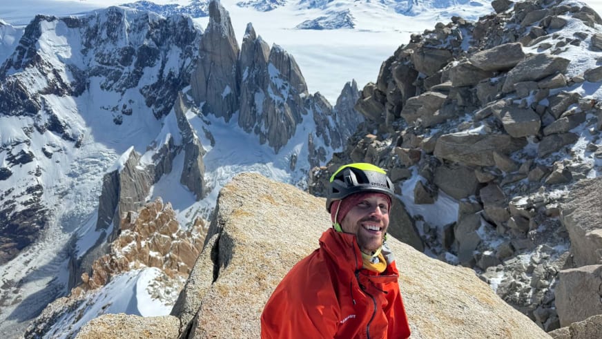 Bergführer Alexander Berger lächelt am Felsgipfel mit Kletterhelm und roter Bergjacke vor Berglandschaft.