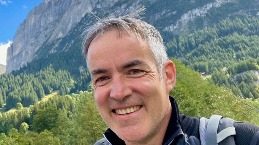 Bergwanderführer Alexander Hoffmann - Kuhnt lächelt in die Kamera, trägt grauen Rucksack vor alpiner Felswand-Kulisse.