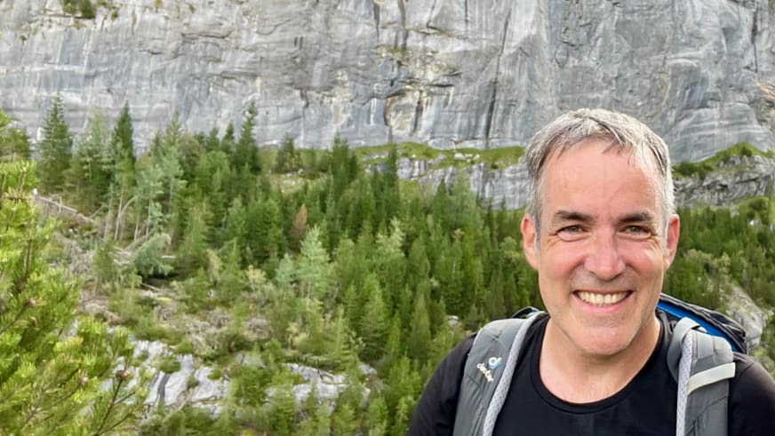 Lächelndes Portrait von Bergwanderführer Alexander Hoffmann - Kuhnt mit schwarzem Rucksack vor einer Kalkstein-Felswand mit Nadelwald.